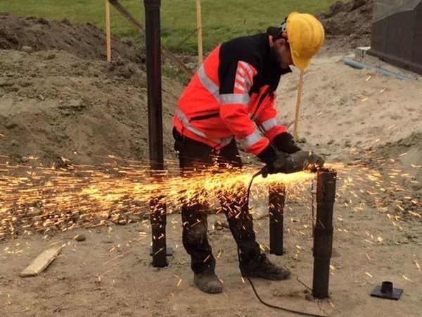 Punaisiin turvavarusteisiin pukeutunut työntekijä leikkaa metalliputkea kipinöiden lentäessä rakennustyömaalla