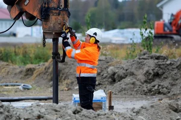 Oranssiin turvavarusteisiin pukeutunut työntekijä käyttää porauslauttaa mutaisella rakennustyömaalla
