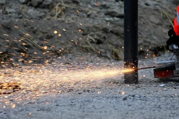 Kulmahiomakone leikkaa mustaa metallipylvästä ja heittää kirkkaan oransseja kipinöitä soraan.