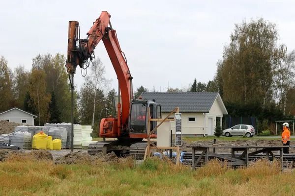 Oranssi kaivinkone rakennustyömaalla, taustalla työntekijöitä, materiaalipinoja ja taloja