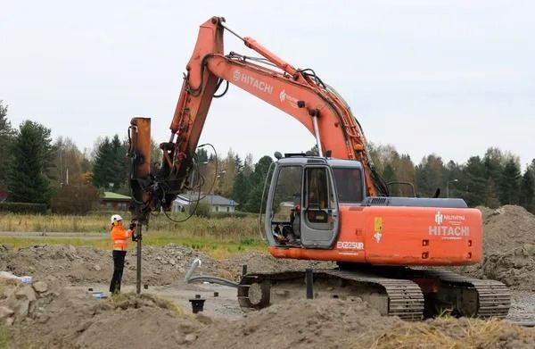 Oranssi kaivinkone poraa maata heijastinliiviin pukeutuneen työntekijän vieressä rakennustyömaalla
