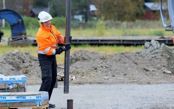 Rakennustyöntekijä oranssissa takissa ja valkoisessa kypärässä käsittelee metallipylvästä työmaalla