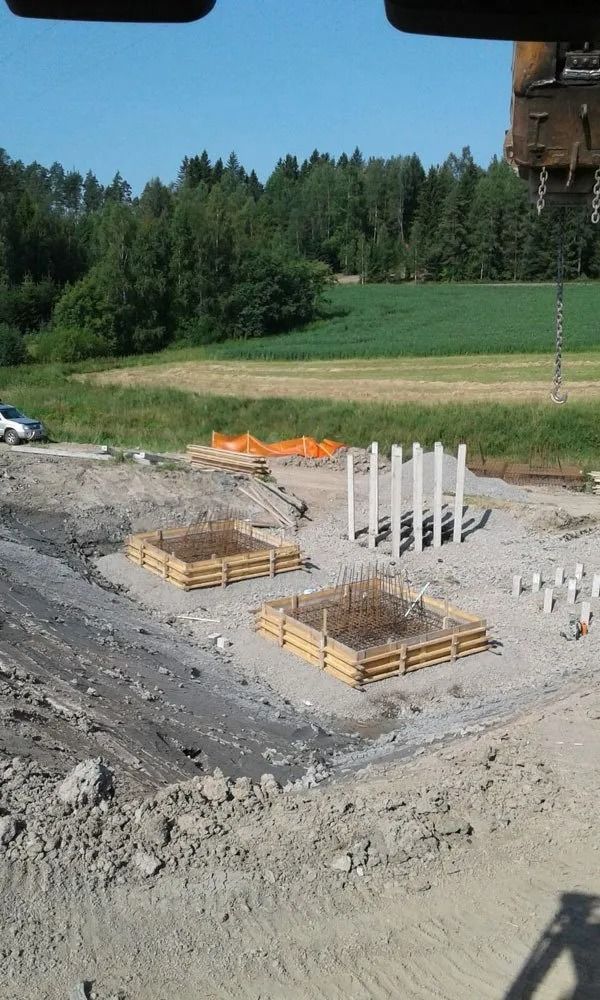 Rakennustyömaa, jossa on betonisia perustusmuotit, raudoitus ja soraa metsäisen rinteen vieressä
