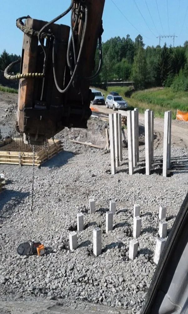Rakennustyömaa, jossa on kaivinkone, sorakasoja ja betonipylväiden pinoja tien lähellä.