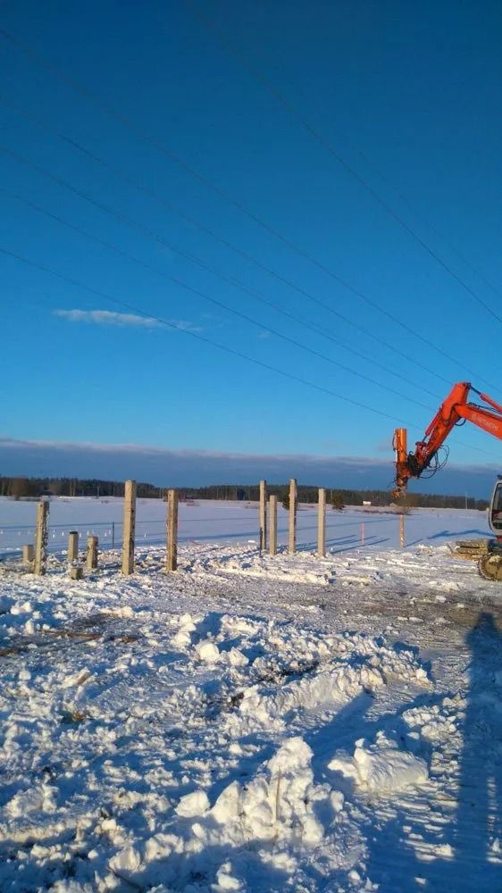 Luminen rakennustyömaa, jossa on puupylväitä ja oranssi kaivinkone kirkkaan sinisen taivaan alla