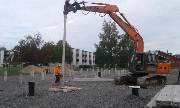 Oranssi kaivinkone nostaa betonipylvästä rakennustyömaalla työntekijän ollessa lähellä
