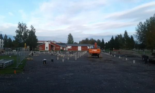 Rakennustyömaa, jossa on oranssi kaivinkone, punaisia rakennuksia ja puita pilvisen taivaan alla