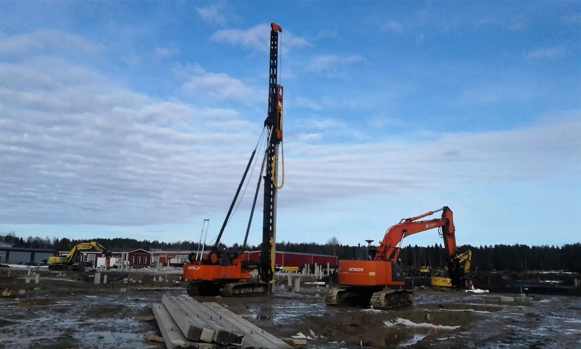Rakennustyömaa, jossa on korkea porauslauta ja kaivinkoneita mutaisella maalla pilvisen taivaan alla
