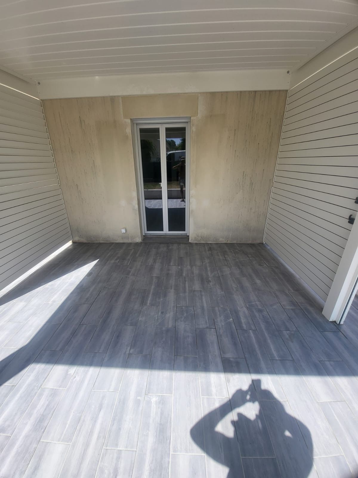 Patio couvert avec sol carrelé gris, côtés à lattes blanches et porte donnant sur l'intérieur. La lumière du soleil projette une ombre.