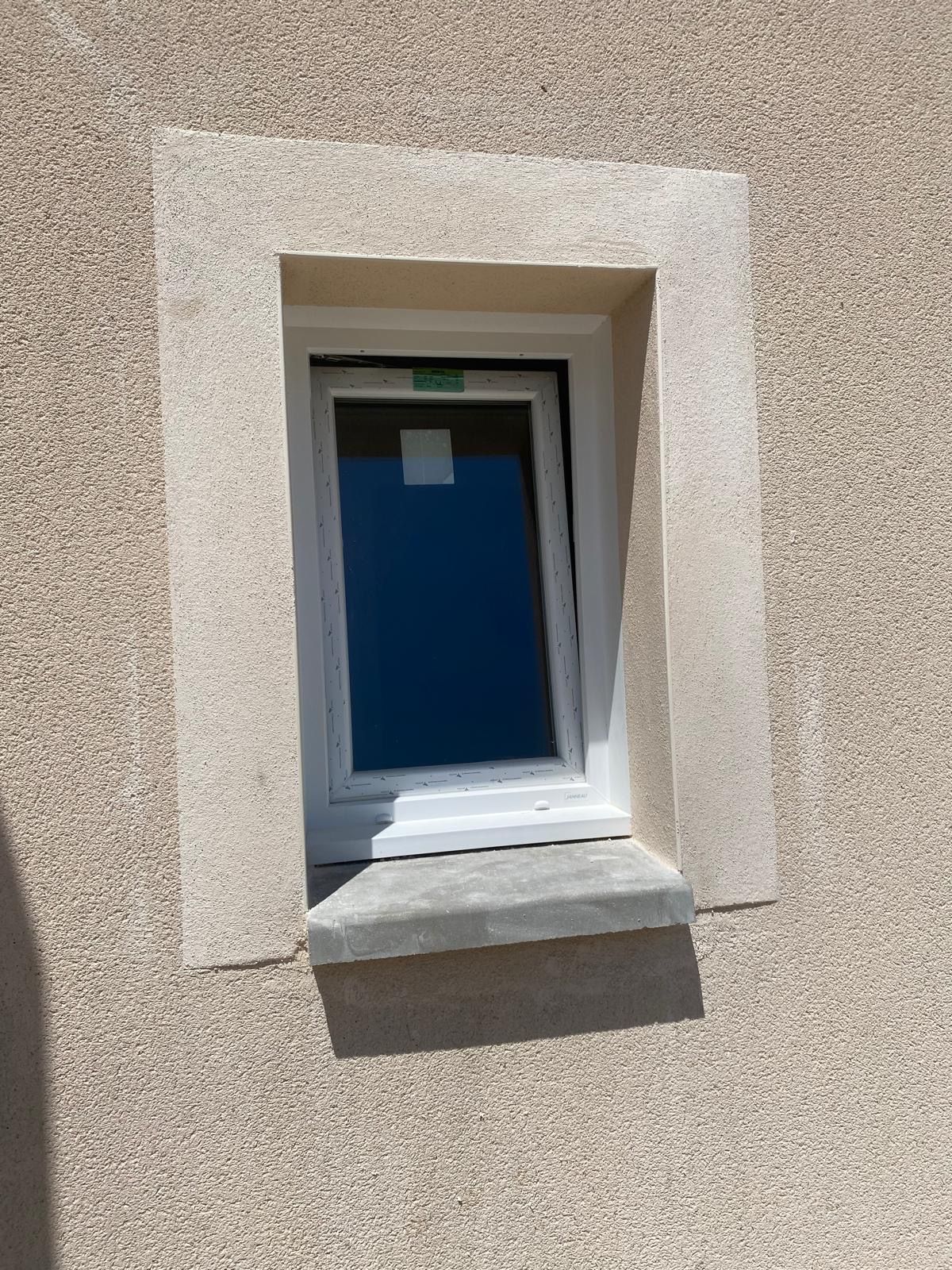 Fenêtre sur mur extérieur, ouverte en biais, avec un appui en pierre et un encadrement en stuc beige.