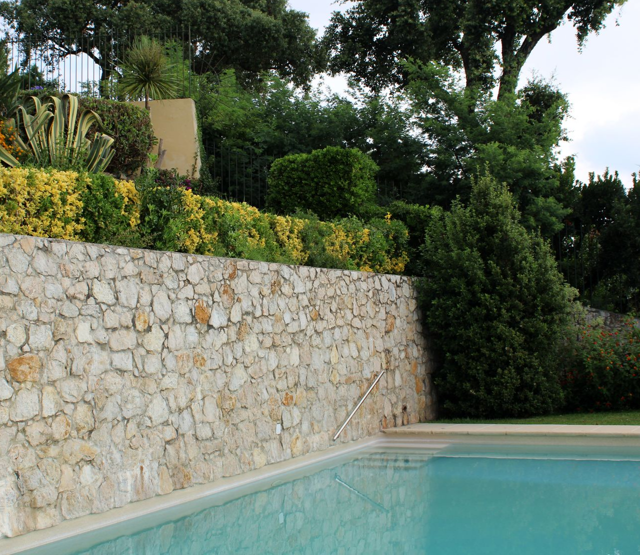 Mur en pierre bordant une piscine, avec de la verdure au-dessus et des arbres en arrière-plan.