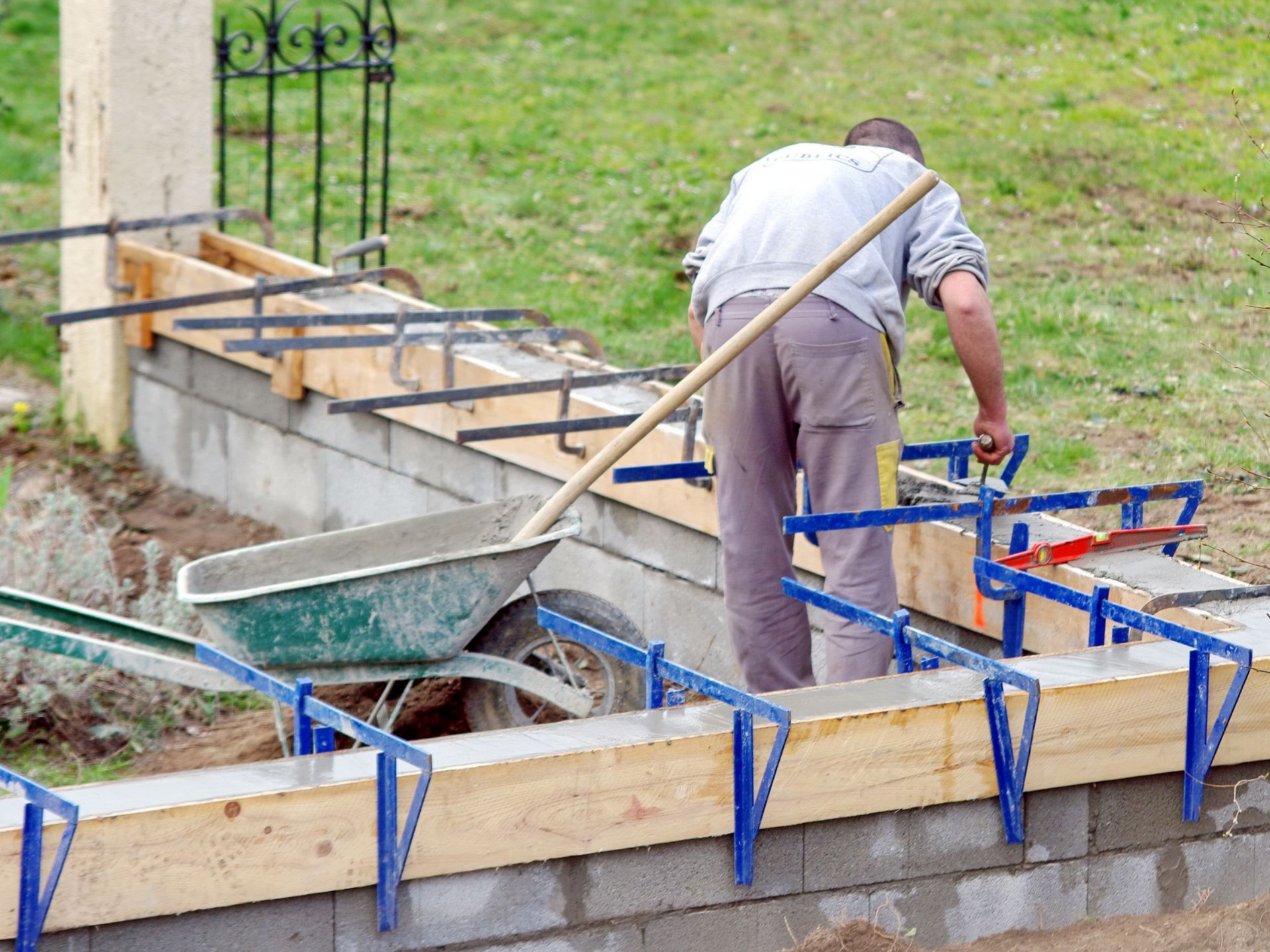 Un ouvrier du bâtiment travaille sur un coffrage en béton, une brouette à proximité.