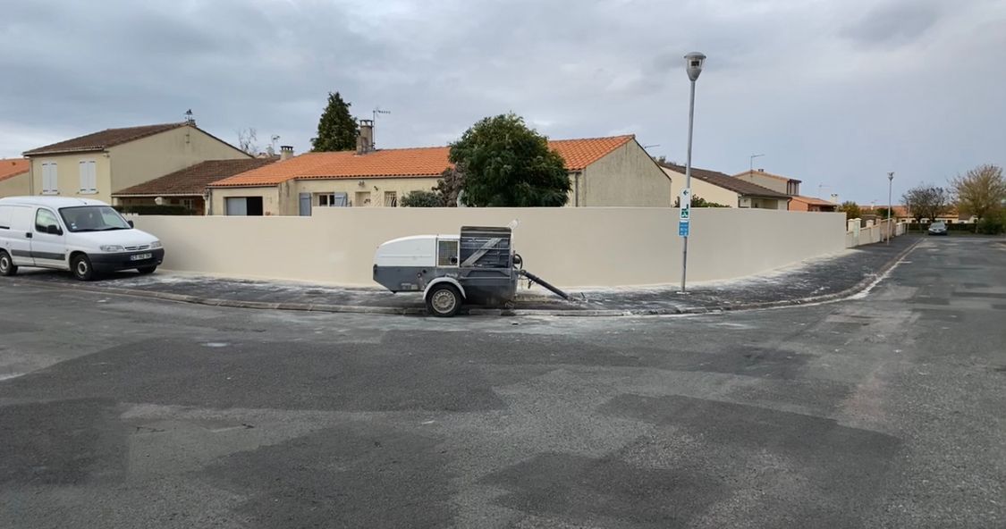 Une camionnette blanche est garée près d'un mur beige fraîchement peint. Une petite remorque et des bâtiments se trouvent à l'arrière-plan.