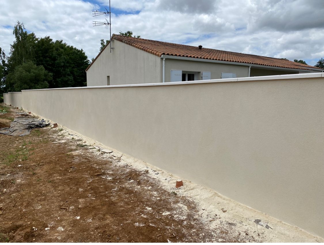 Mur en stuc beige jouxtant un chemin de terre, une maison au toit de tuiles rouges se dresse en arrière-plan.
