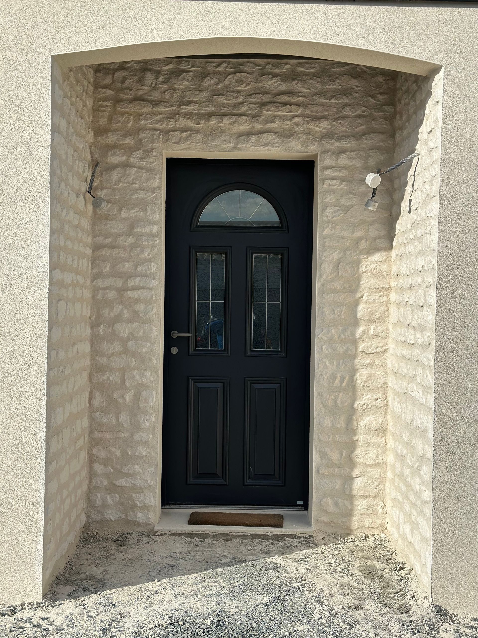 Porte d'entrée noire avec fenêtre cintrée, située dans une alcôve aux murs beiges texturés, petit espace gravillonné.