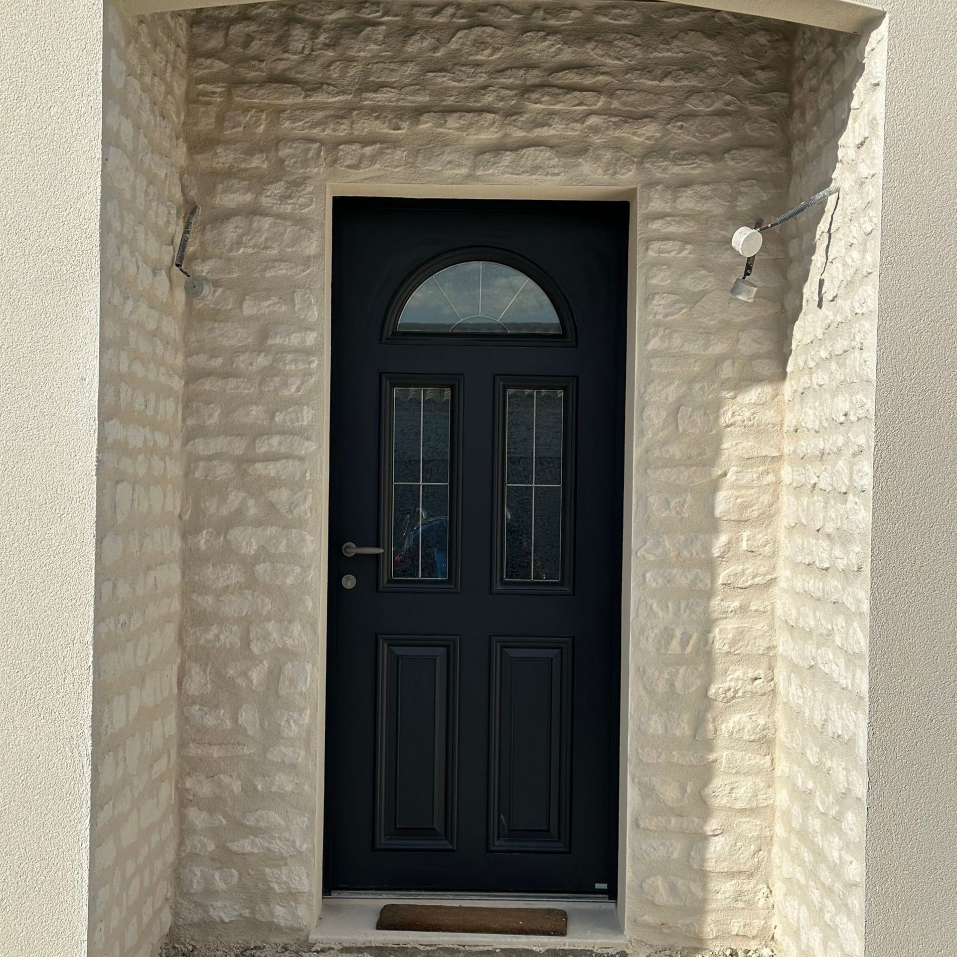 Porte d'entrée noire avec fenêtre cintrée et imposte latérale, encastrée dans une façade en pierre.
