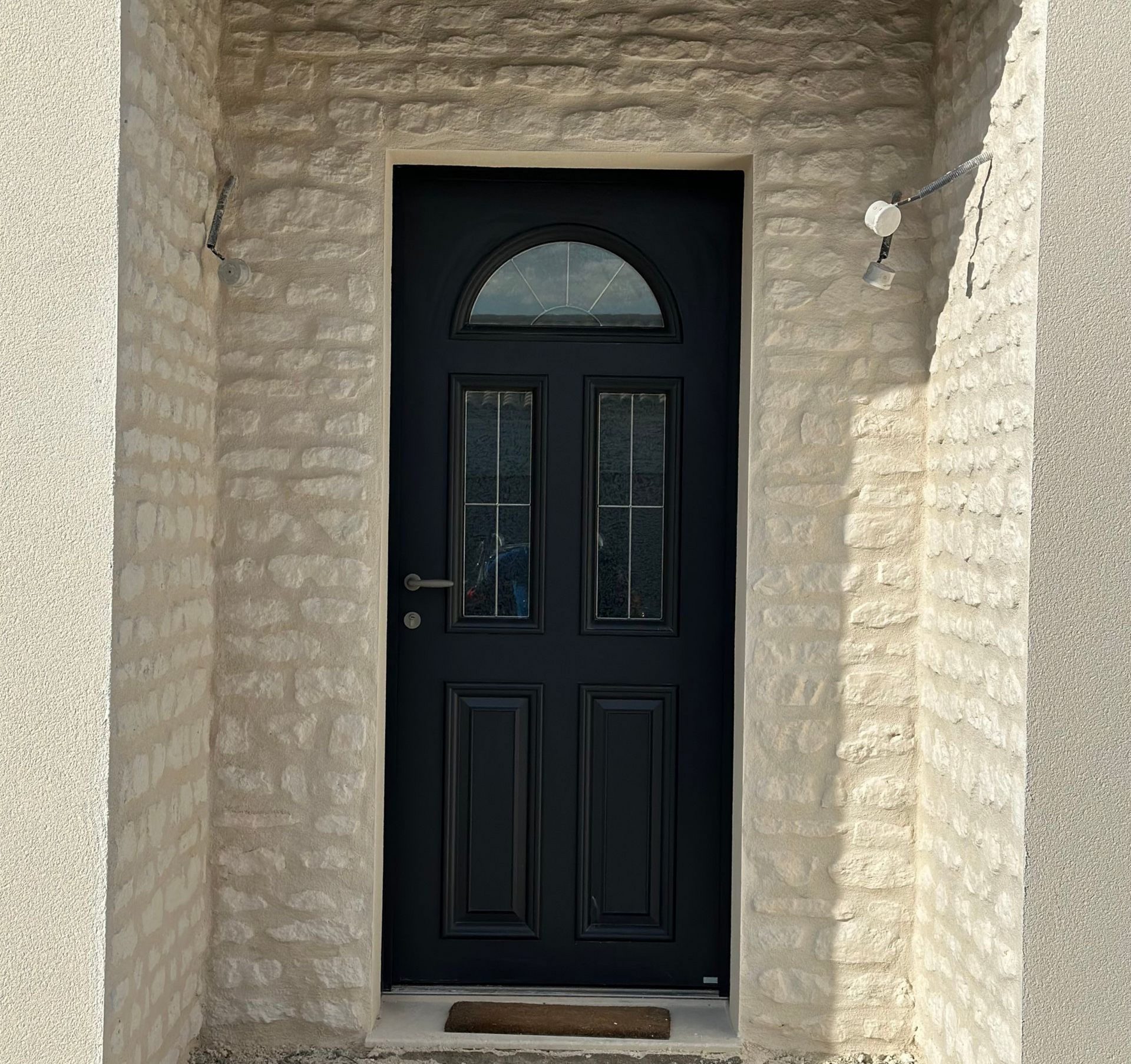 Porte noire vitrée encadrée d'une arche en pierre. Un petit tapis est posé au pied de la porte.