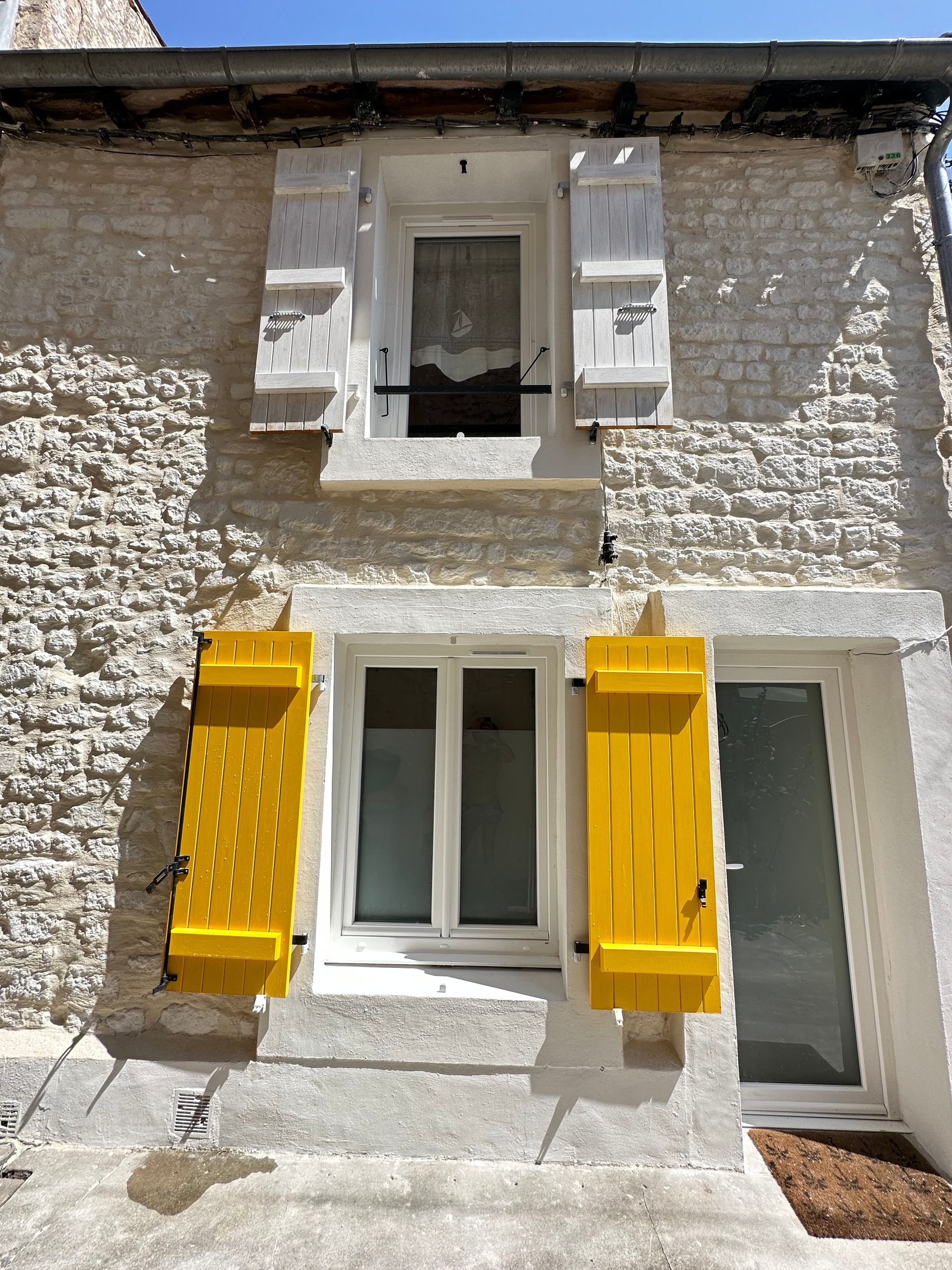 Bâtiment en pierre avec une fenêtre et des volets jaune vif au rez-de-chaussée, des volets blancs à l'étage.