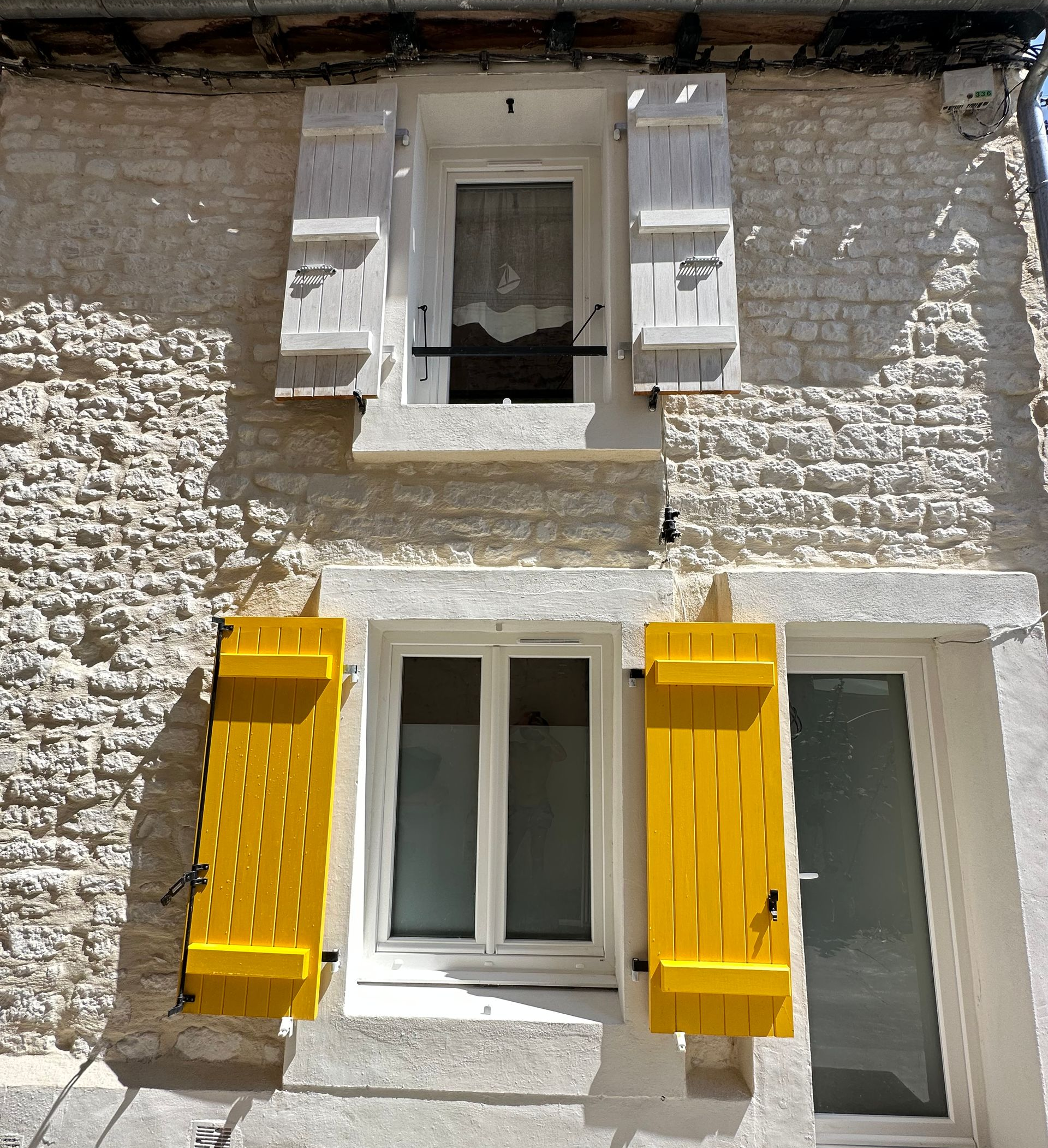 Bâtiment en pierre de deux étages avec des volets jaunes et blancs. La fenêtre du haut a des volets blancs, celle du bas des volets jaunes.