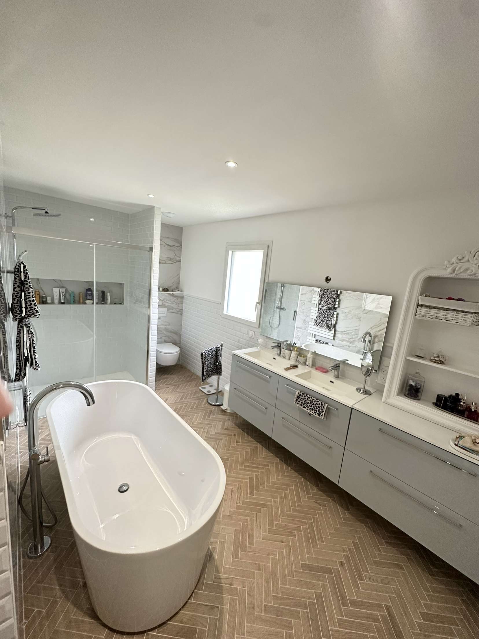 Salle de bains moderne avec baignoire îlot, douche en verre, double vasque et sol carrelé clair.