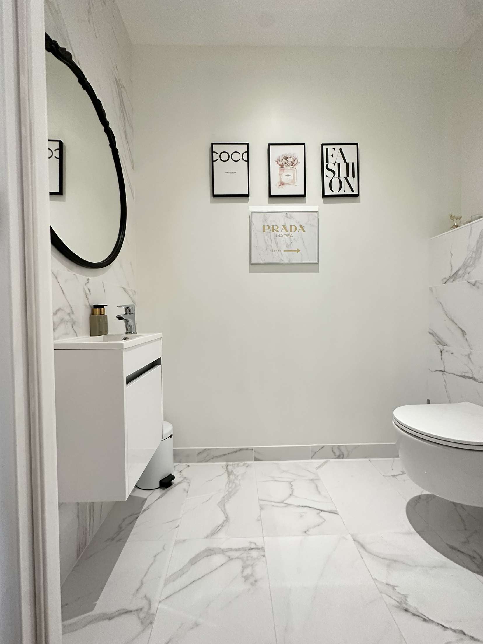 Salle de bains moderne blanche avec sol en marbre, meuble-lavabo suspendu, miroir ovale et trois estampes encadrées.