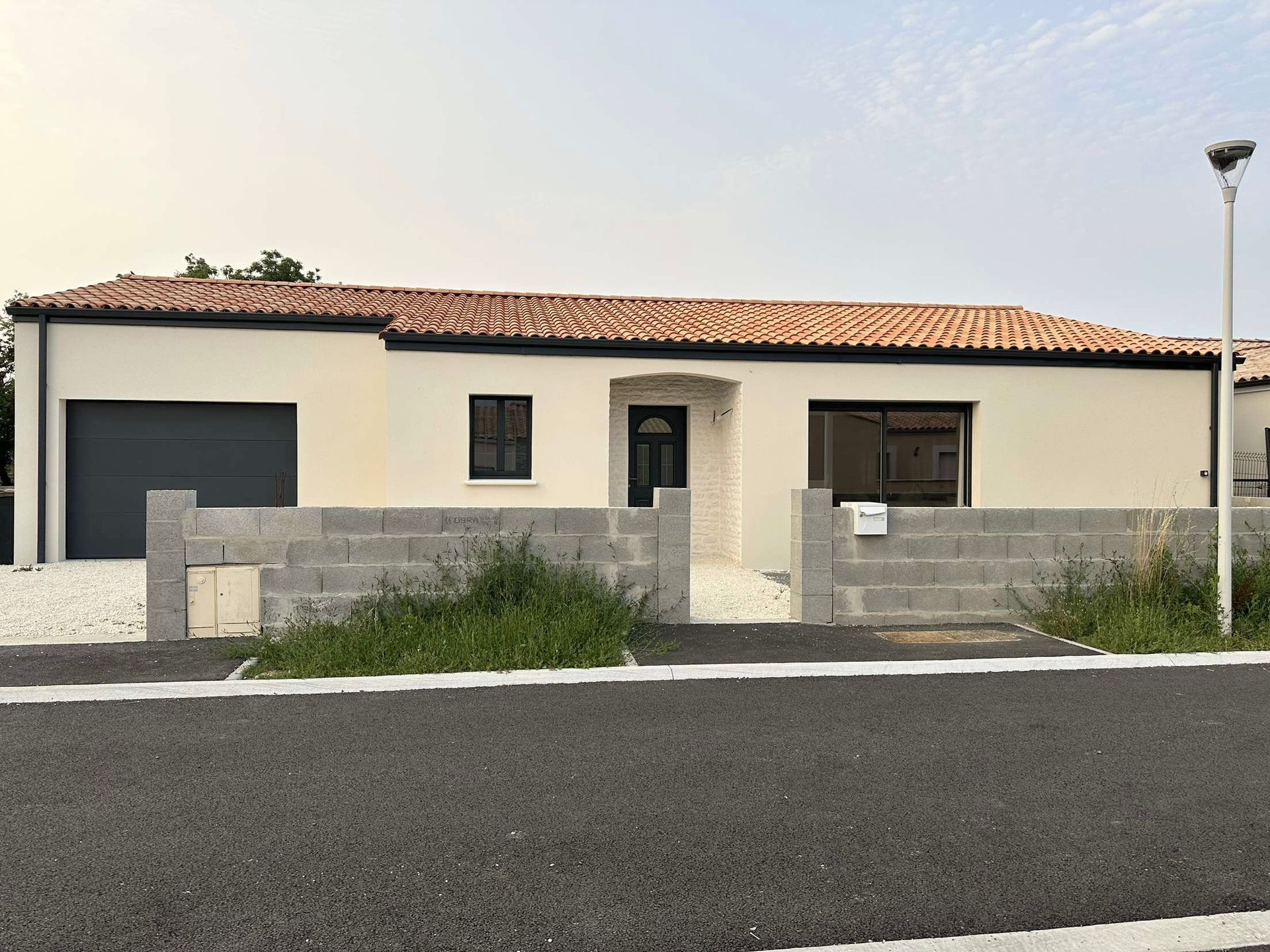 Maison beige avec un toit de tuiles rouges, une porte de garage grise et une clôture en parpaings de béton, située dans une rue pavée.