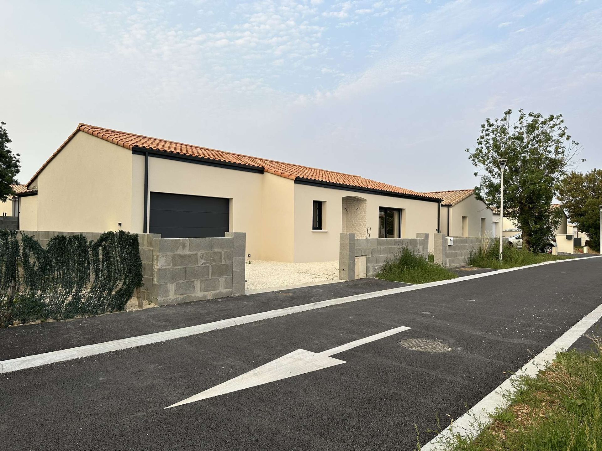 Maison beige avec toit de tuiles rouges, clôture en parpaings, route asphaltée avec flèche peinte.