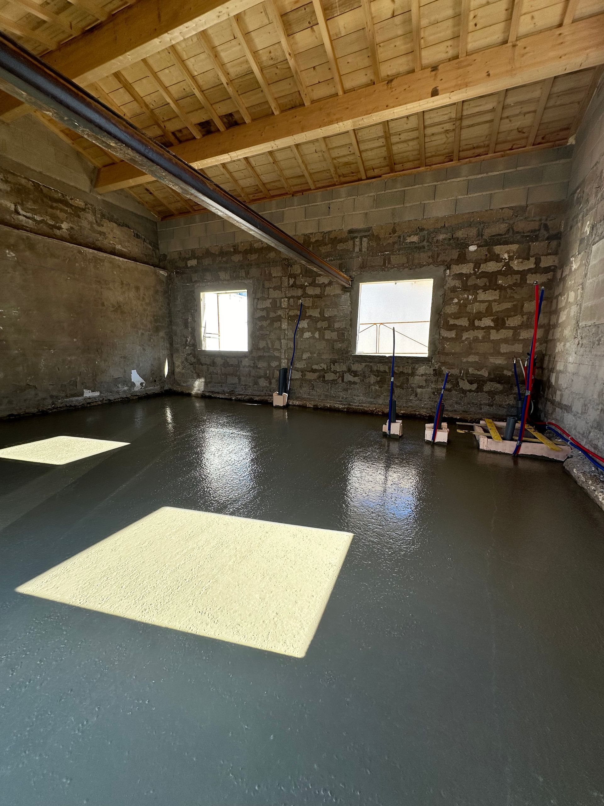 Pièce intérieure avec sol en béton frais et humide ; murs en pierre ; poutres apparentes. Deux fenêtres.