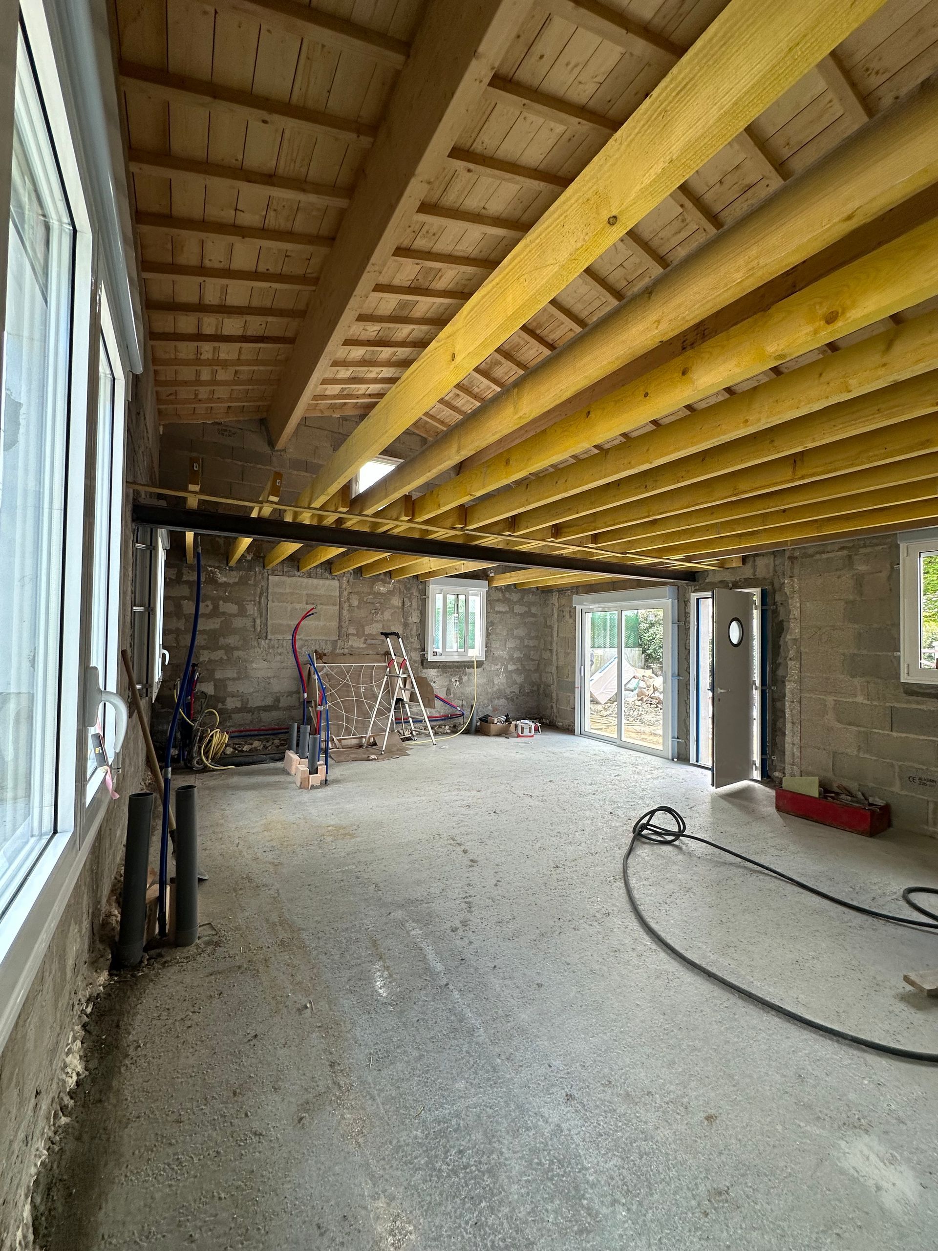 Sous-sol non aménagé avec poutres apparentes, sol en béton et grandes fenêtres. Outils et matériaux de construction présents.
