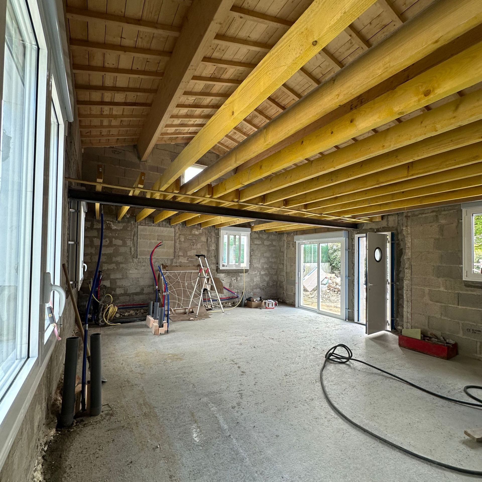 Intérieur d'une pièce en construction avec poutres en bois apparentes, sol en béton, fenêtres et porte.