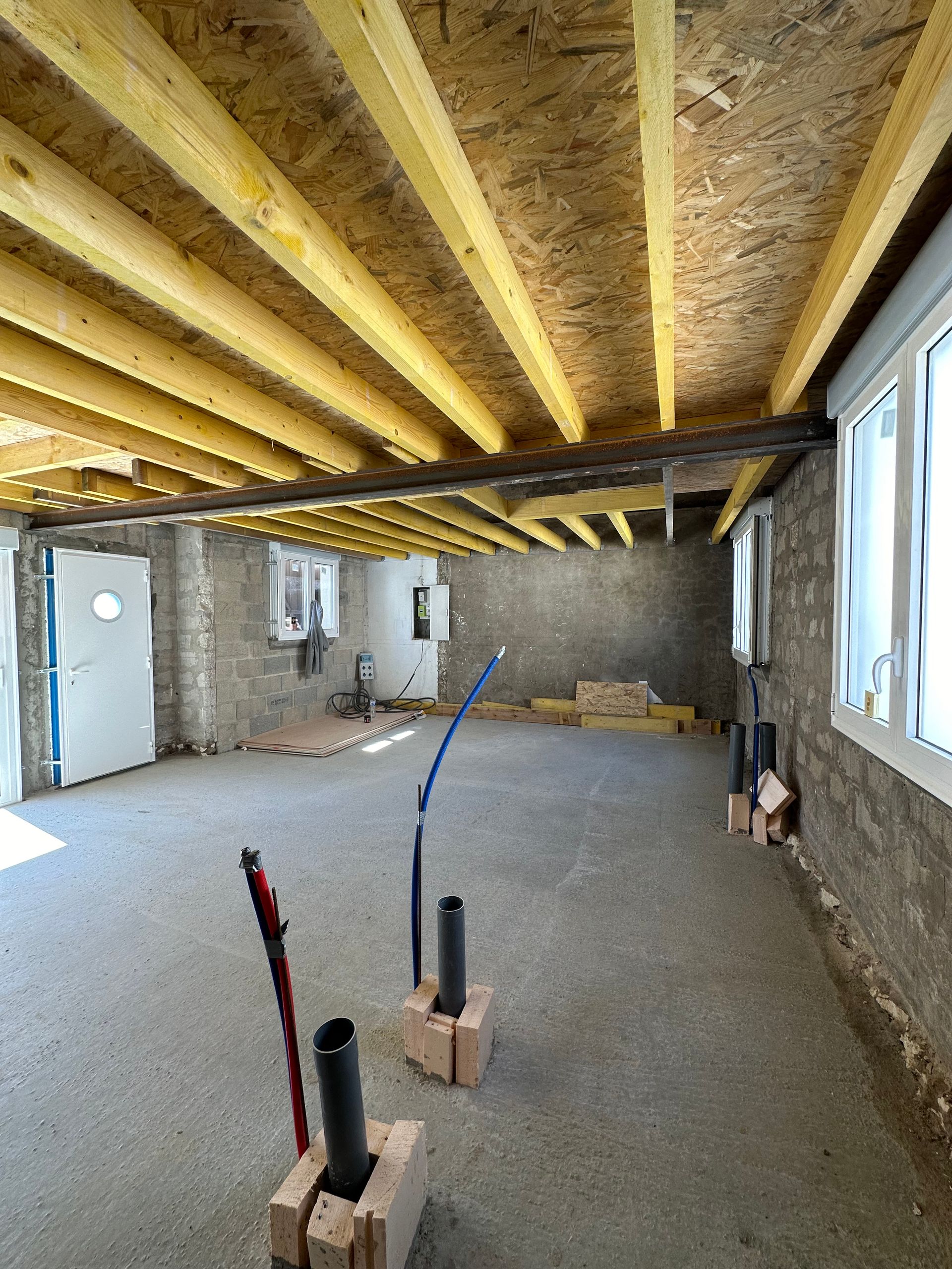 Intérieur d'une pièce inachevée ; poutres en bois apparentes, sol en béton, murs en briques et tuyauterie visible.