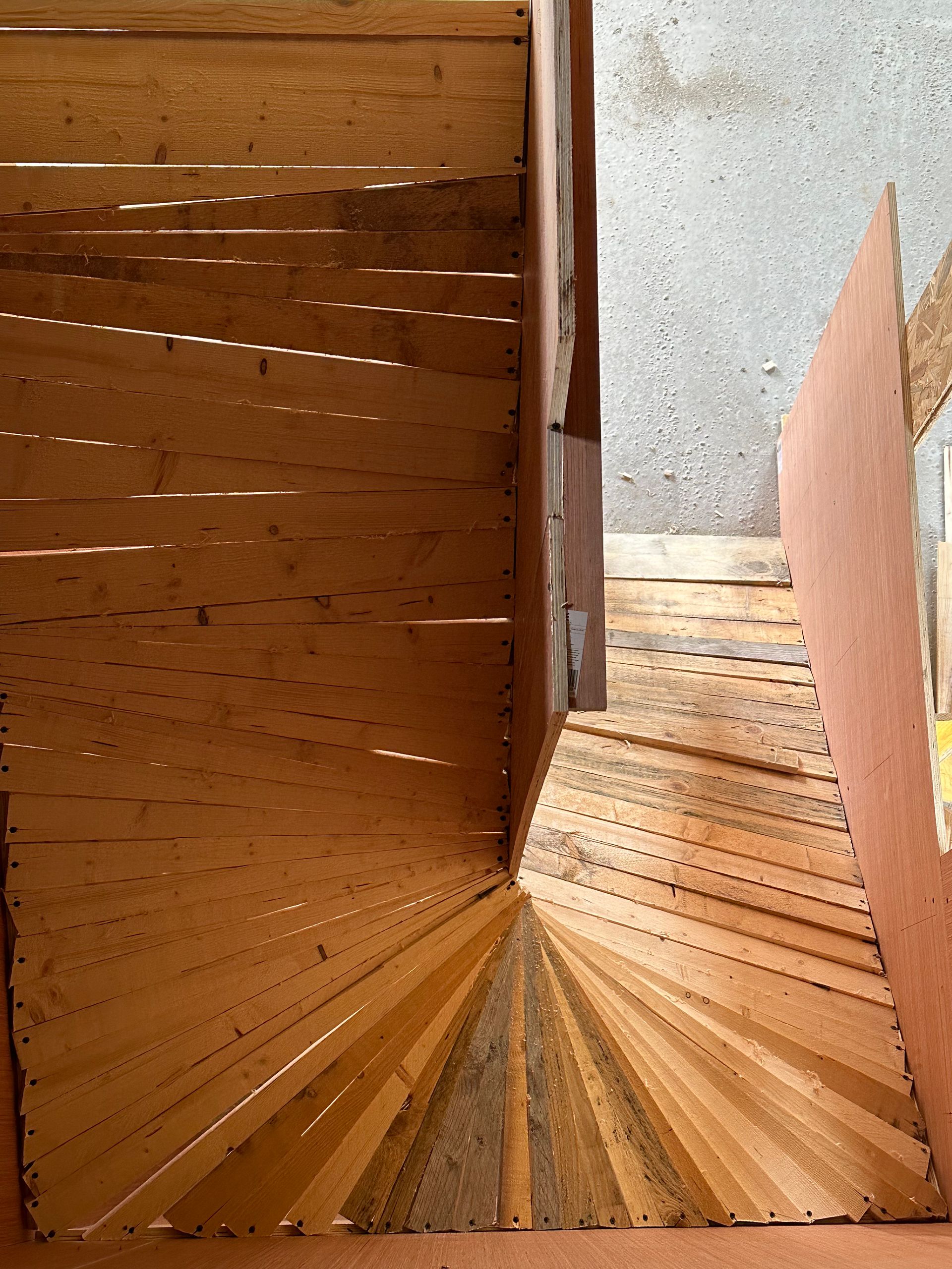 Escalier en bois à marches obliques menant à un virage. On aperçoit du bois brun et un mur en béton.