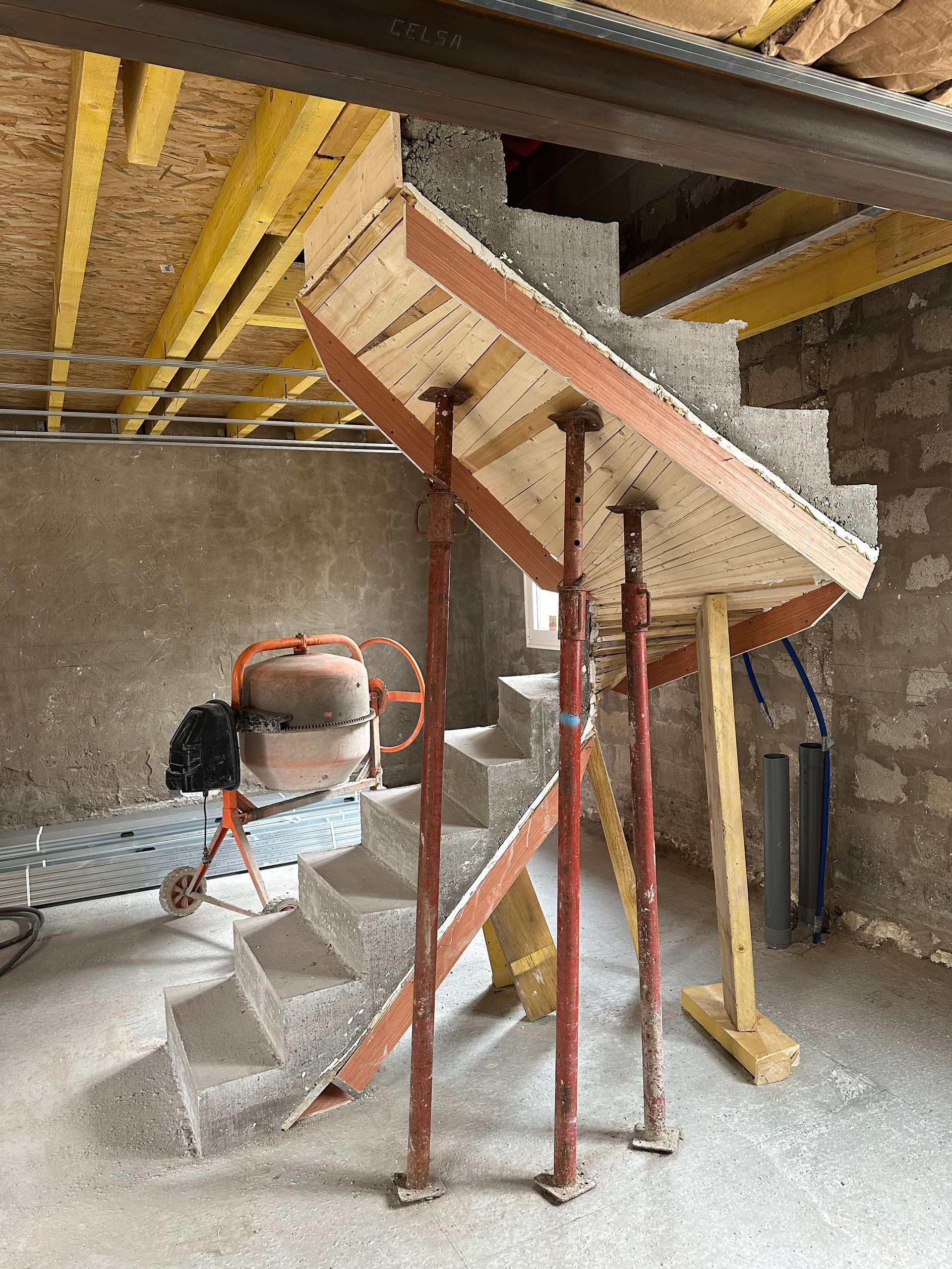 Escalier en construction avec marches en béton et coffrage en bois soutenu par un échafaudage. Bétonnière en arrière-plan.