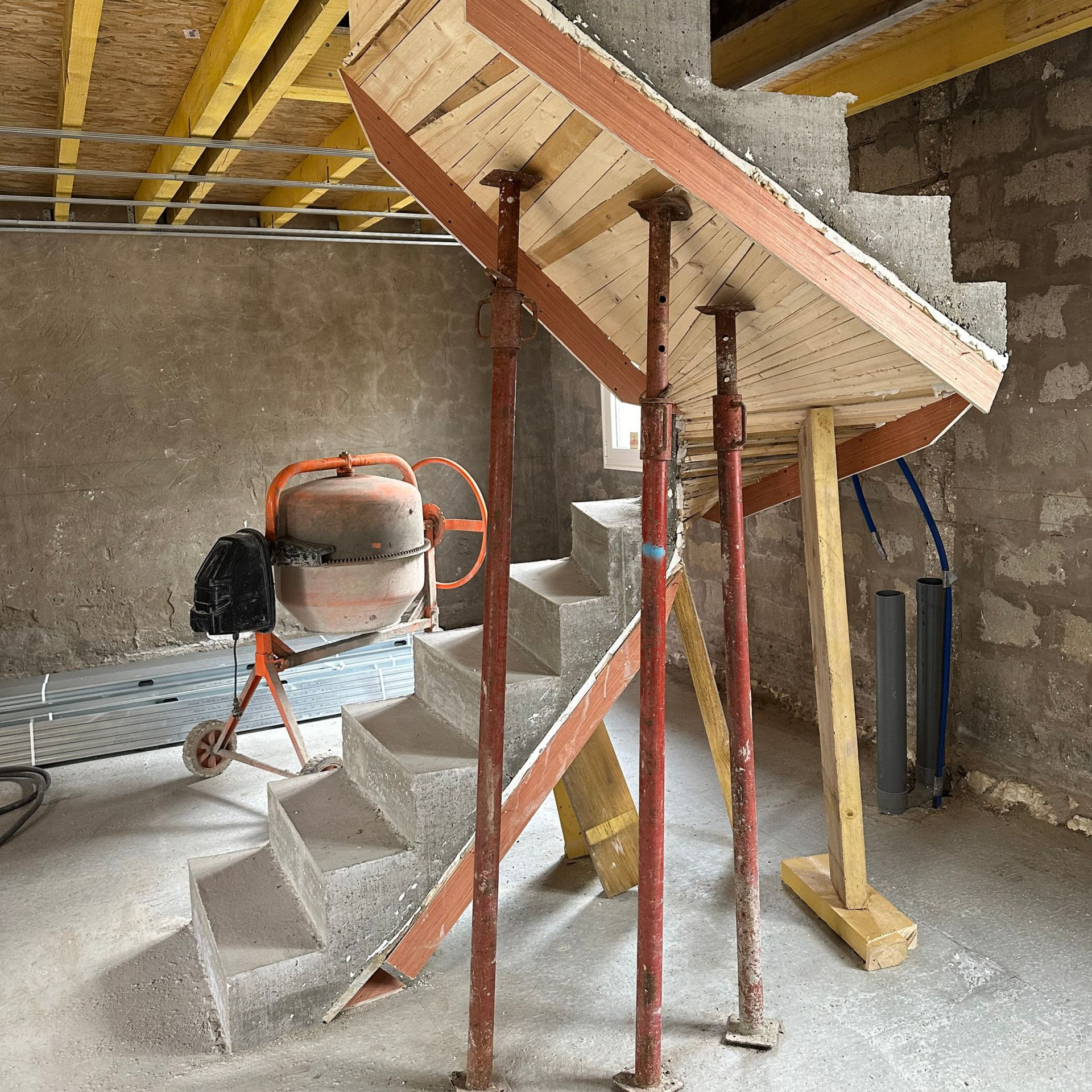 Chantier de construction avec escalier en béton et structure d'escalier en bois soutenue par des poteaux métalliques ; bétonnière en arrière-plan.