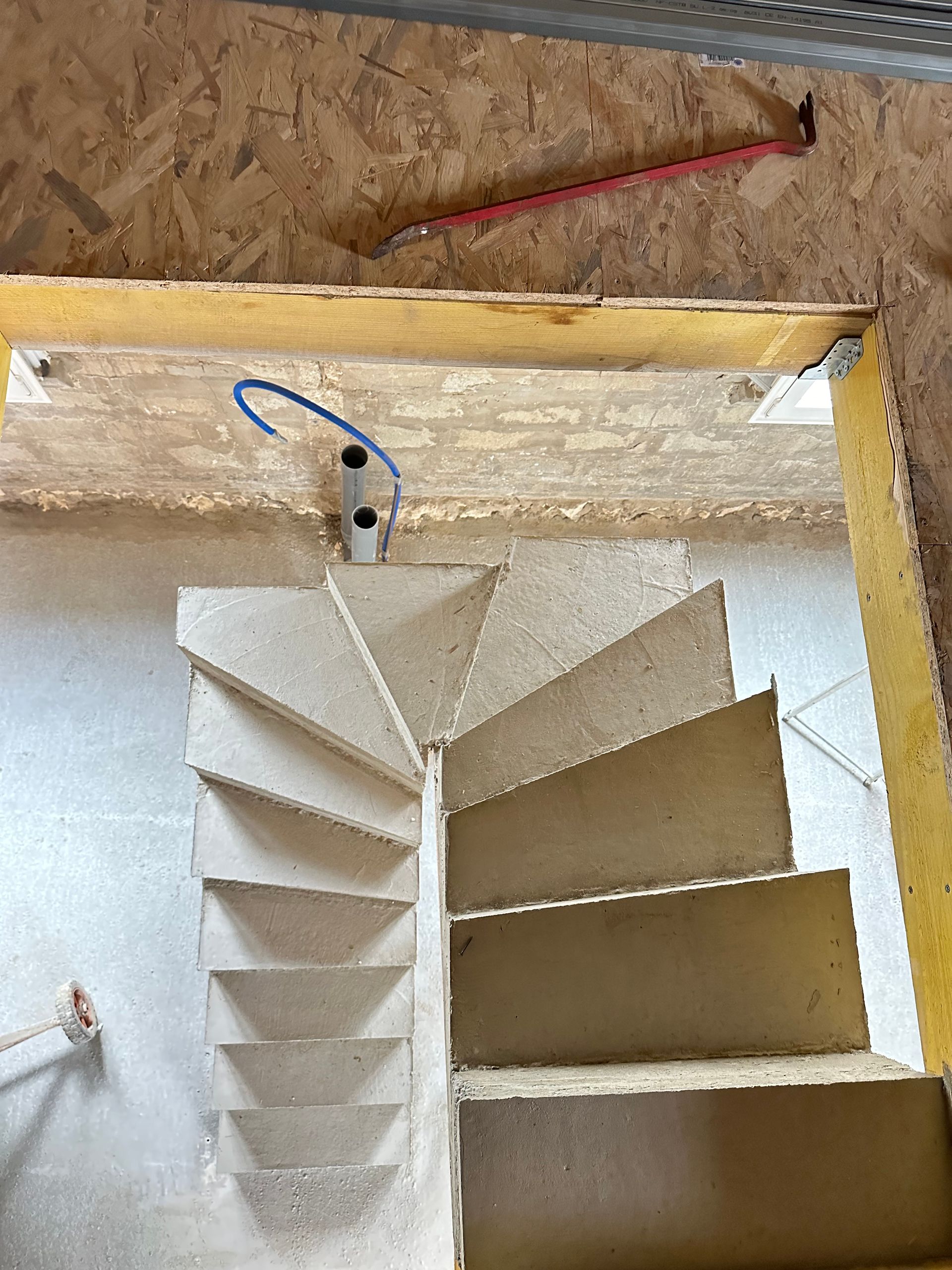 Escalier en colimaçon en construction ; bois apparent, marches en béton, tuyau bleu, fil rouge.