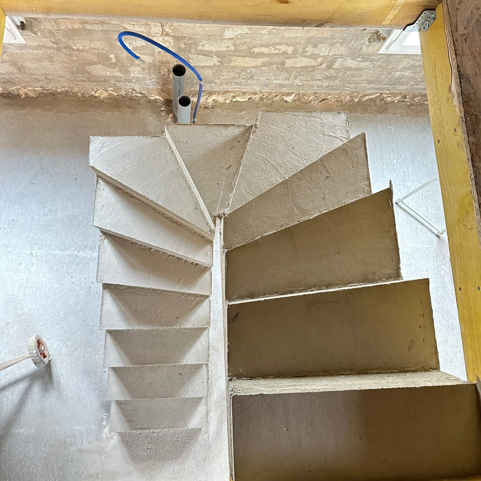 Escalier en colimaçon en construction ; marches beiges, murs en plâtre inachevés, supports en bois.