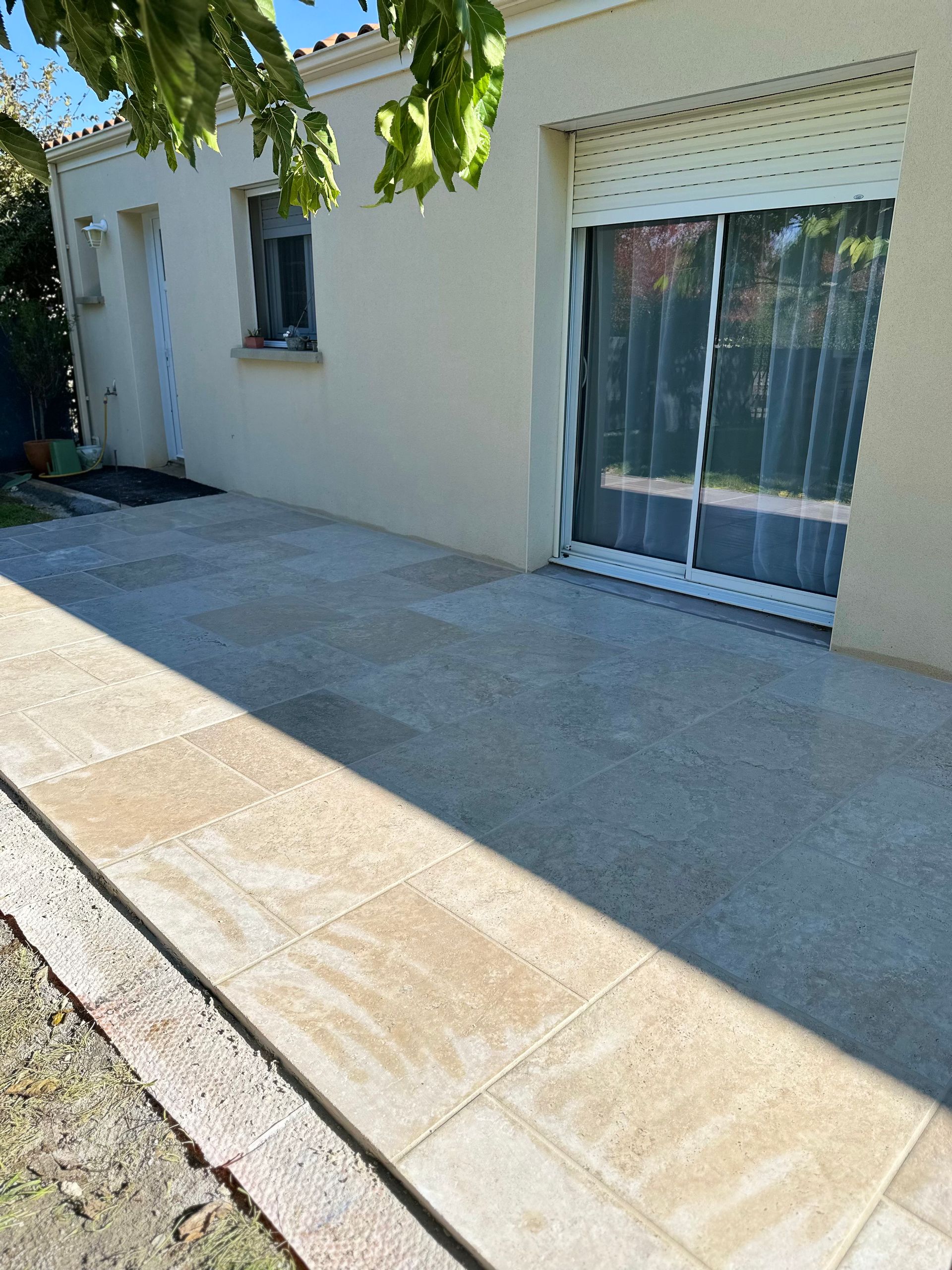 Dalles de terrasse beiges longeant un bâtiment couleur crème avec une fenêtre et une porte coulissante en verre.