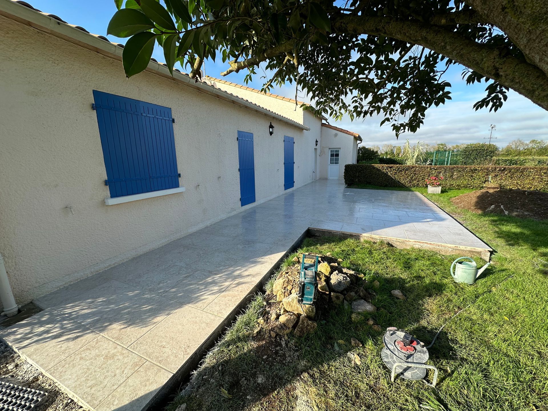 Vue extérieure d'une maison avec des volets bleus, une terrasse en béton et un jardin gazonné sous un arbre.