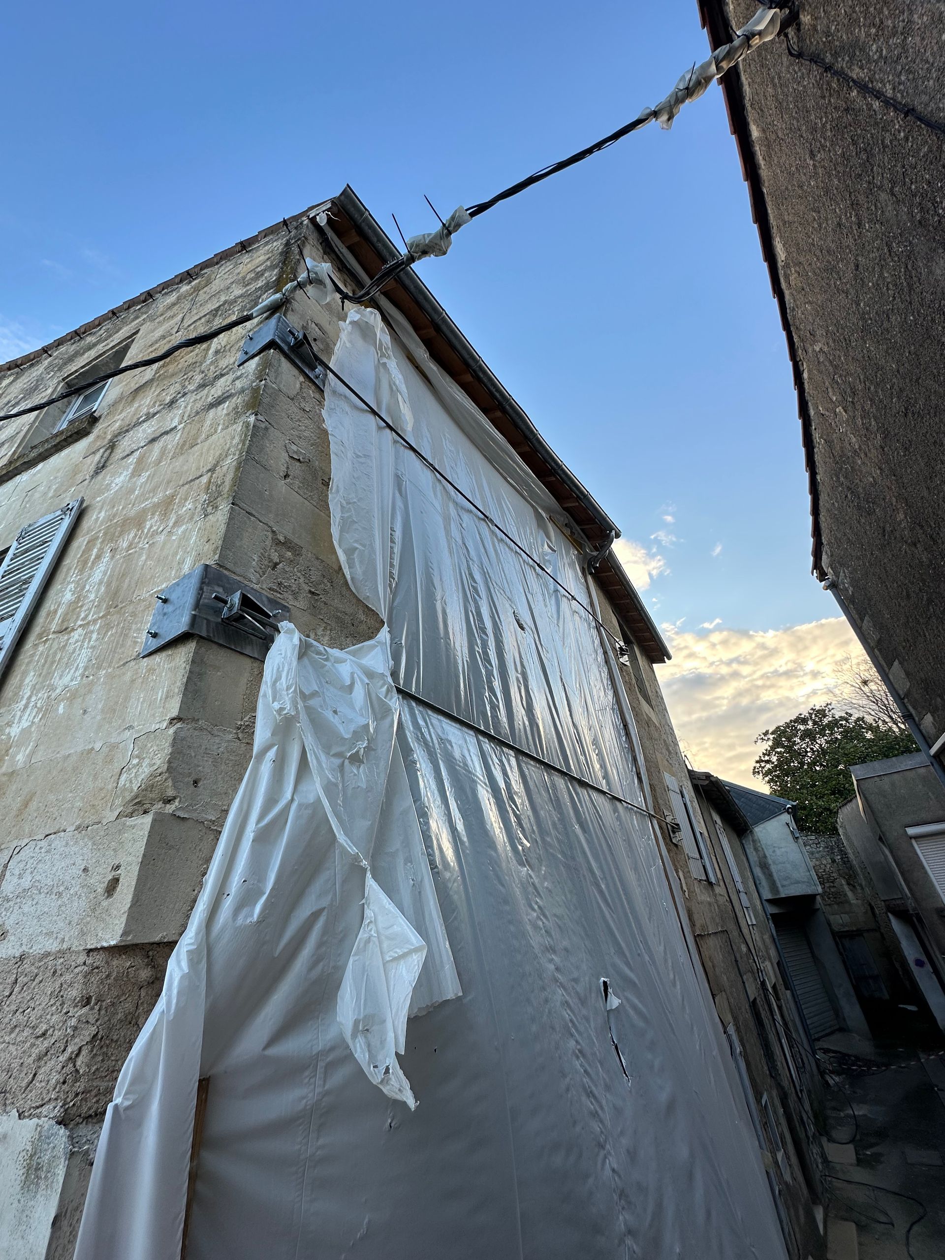 Mur extérieur d'un bâtiment avec un revêtement blanc qui s'écaille ; lignes électriques au-dessus, ciel bleu.