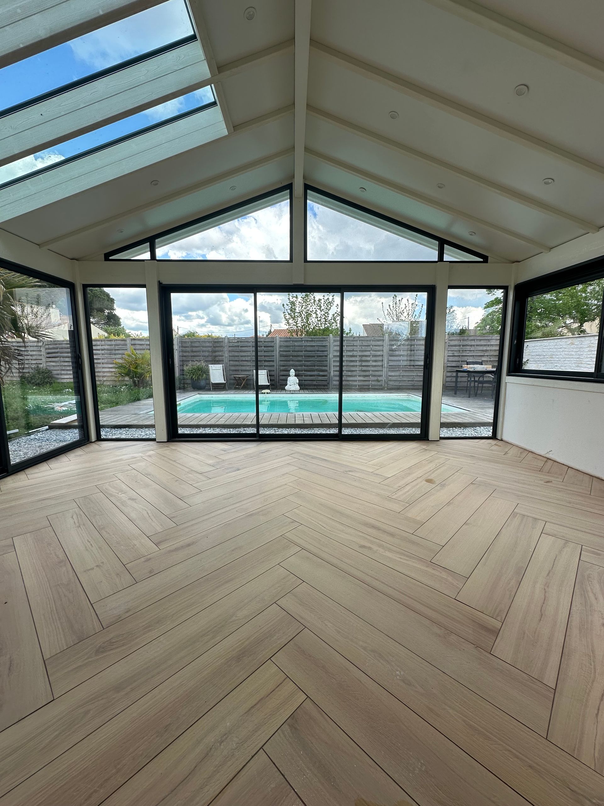 Intérieur d'une véranda avec parquet en chevrons, grandes fenêtres donnant sur une piscine. Ciel visible à travers un puits de lumière.