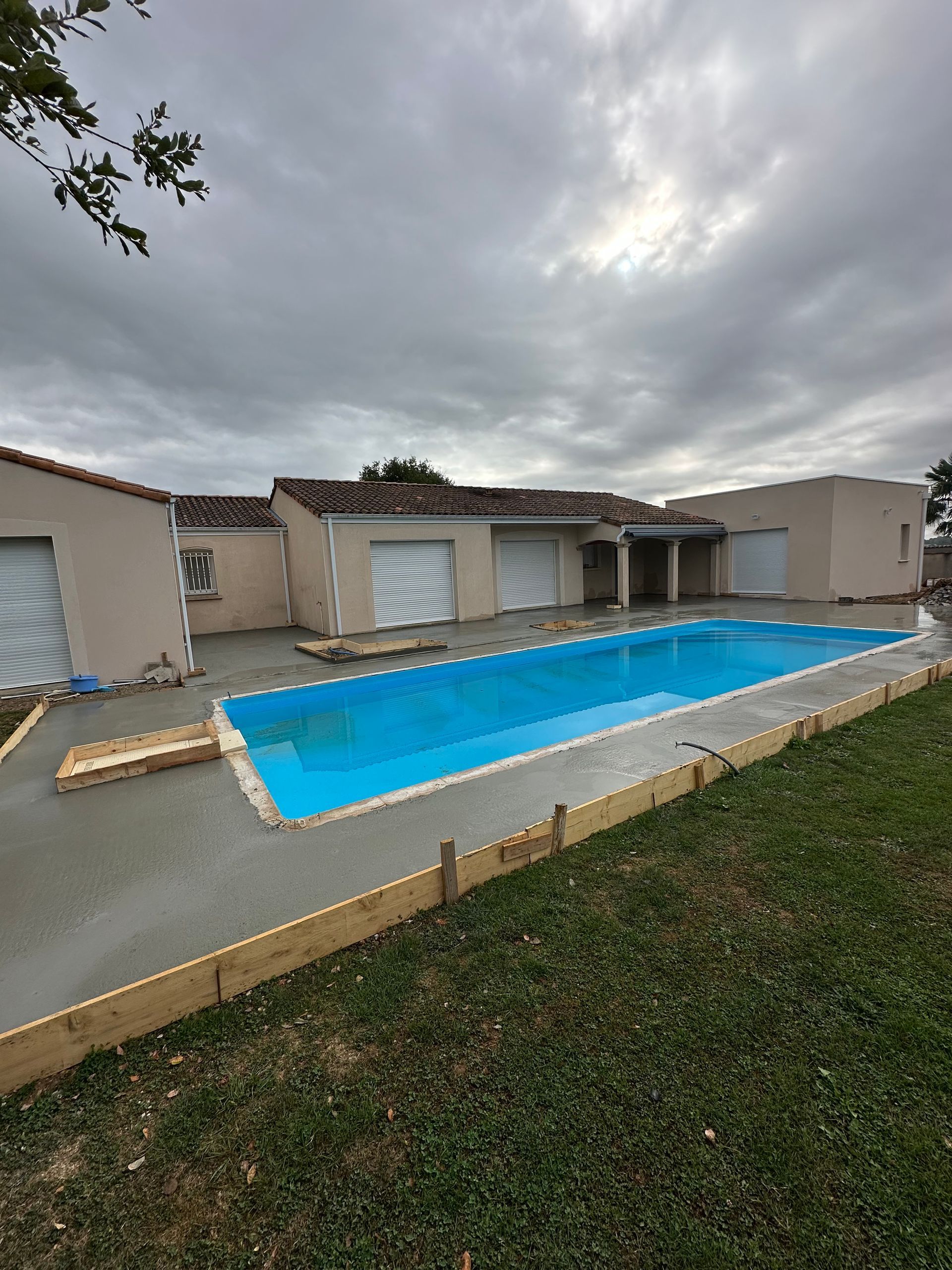Un bassin rectangulaire d'eau bleue se trouve devant une maison aux couleurs claires, sous un ciel nuageux.