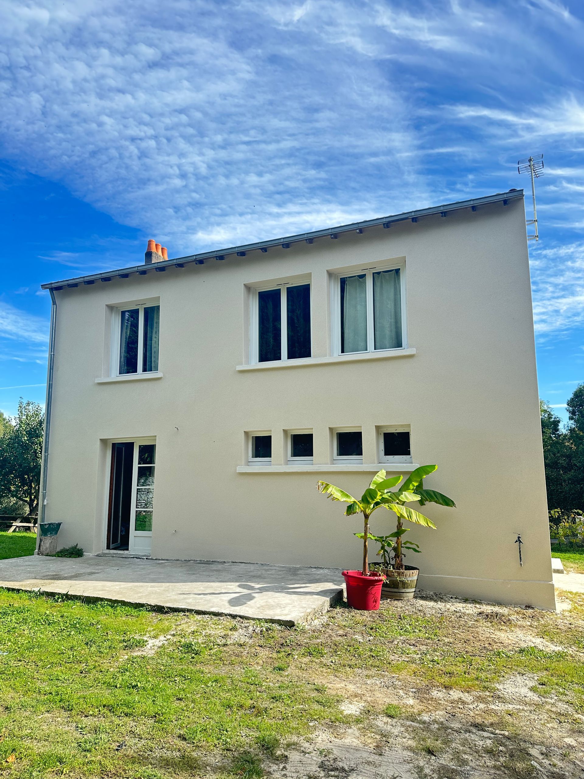 Maison beige à deux étages avec toit en pente, fenêtres et porte ouverte. Petite plante devant, ciel bleu en arrière-plan.