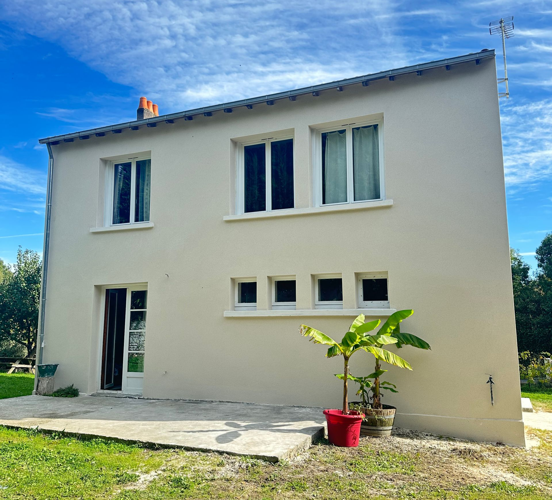 Maison beige à deux étages avec fenêtres et porte blanches, terrasse en béton, jardin verdoyant et ciel bleu.