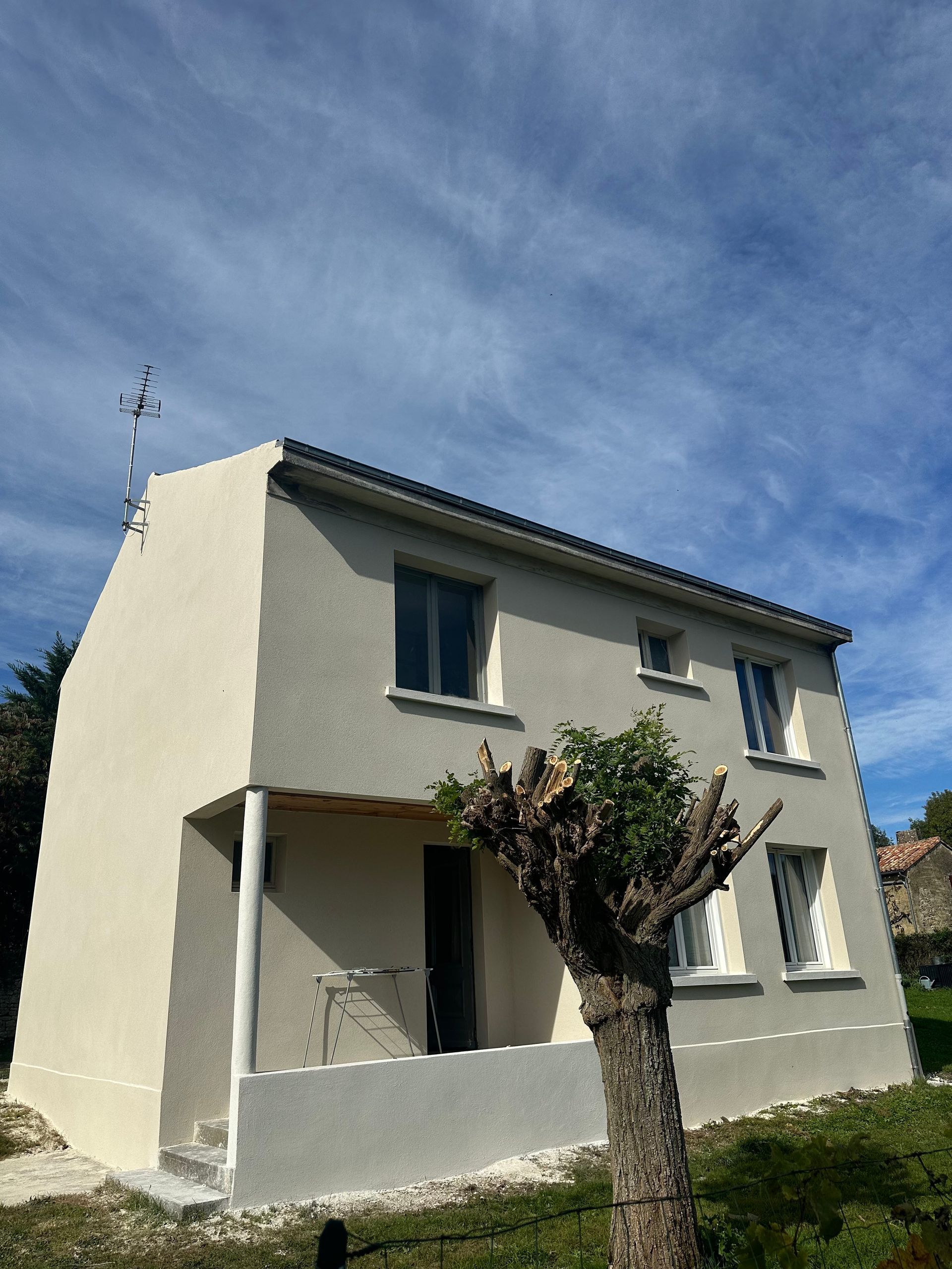 Maison à deux étages avec façade grise et beige, fenêtres et petite véranda sous un ciel bleu.