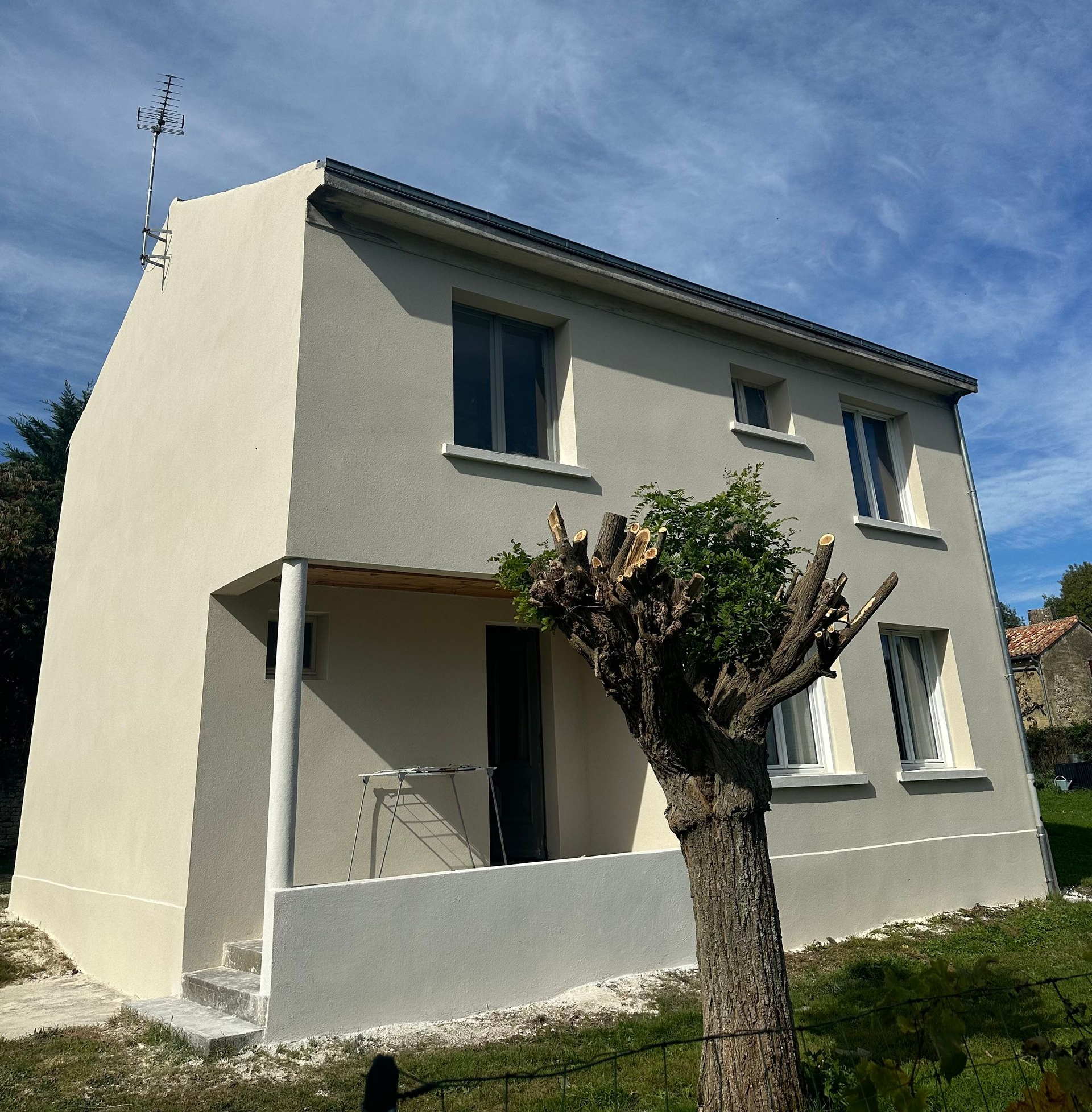 Maison à deux étages avec façade beige clair, fenêtres, petit porche et tronc d'arbre devant le bâtiment.