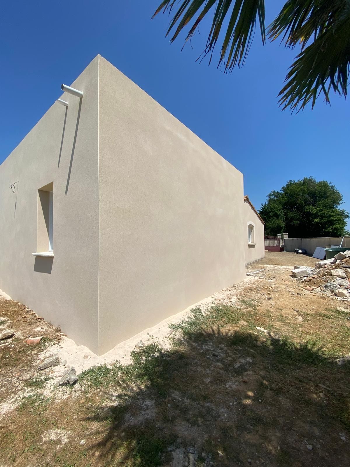 Bâtiment couleur crème avec de petites fenêtres se détachant sur un ciel bleu, une palme dans un coin.