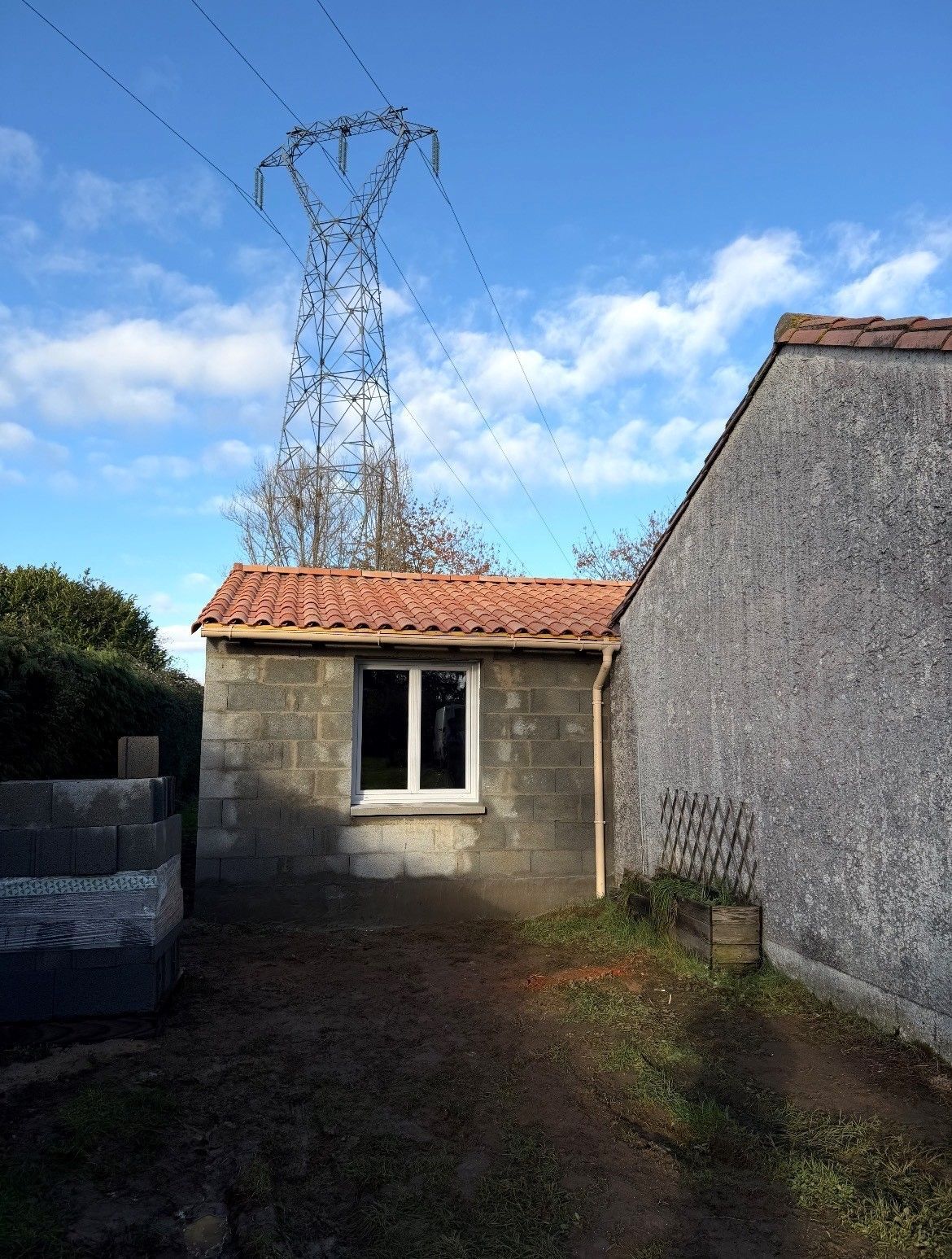 Petit bâtiment en béton avec toit de tuiles, jouxtant une structure grise plus imposante sous une ligne électrique.
