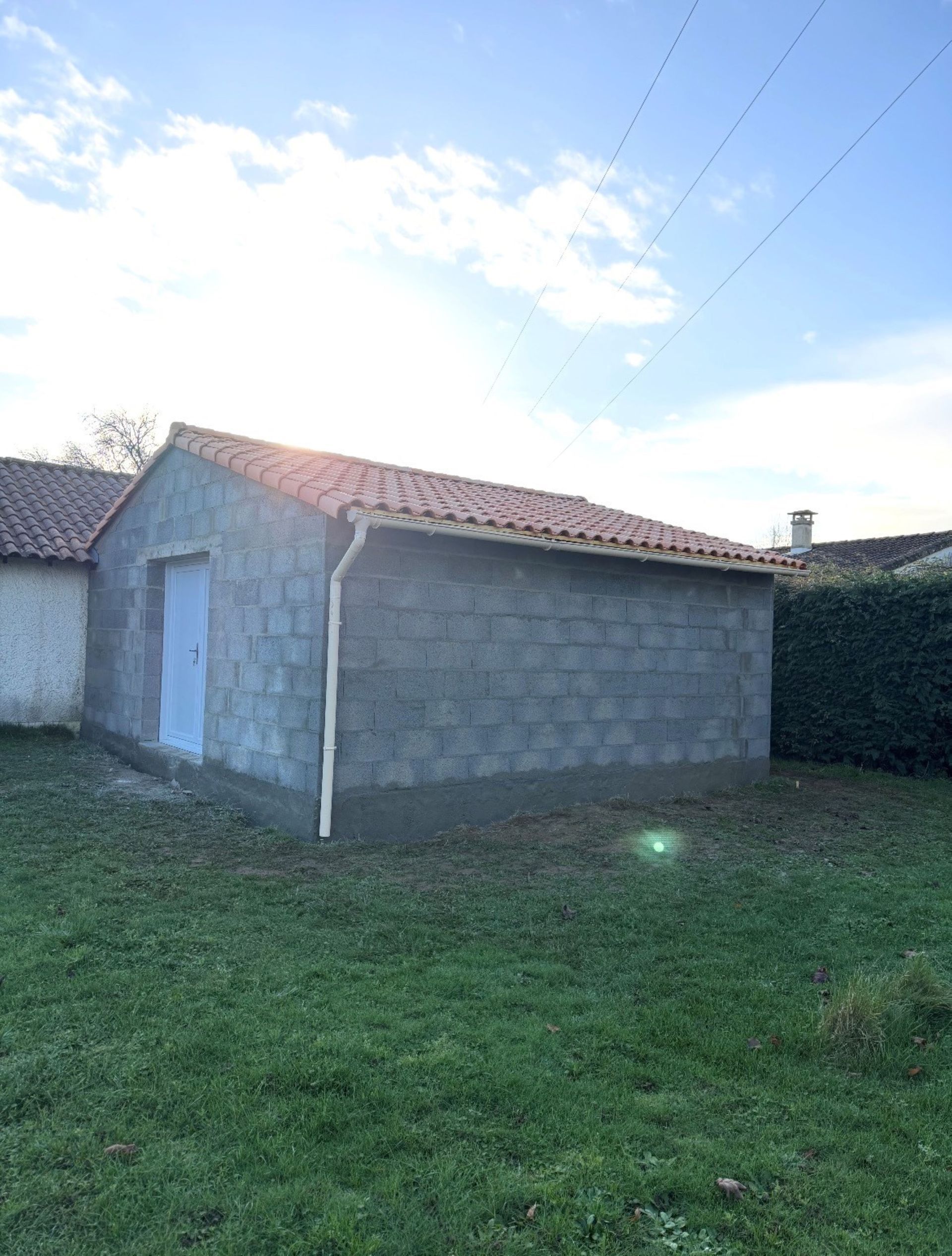 Abri de jardin en parpaings gris avec porte bleue et toit de tuiles rouges, dans une cour arrière gazonnée.