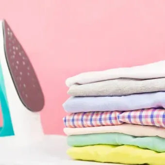 Plancha junto a una pila de ropa doblada, sobre un fondo rosa. La ropa es de varios colores y está perfectamente apilada.