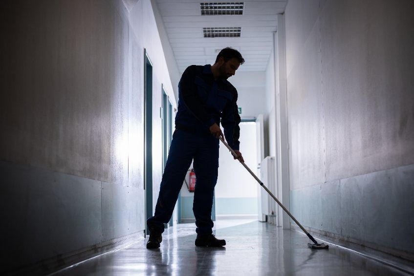 Un hombre vestido con ropa de trabajo oscura friega un suelo brillante en un pasillo largo y sombreado, con paredes blancas y puertas cerradas.